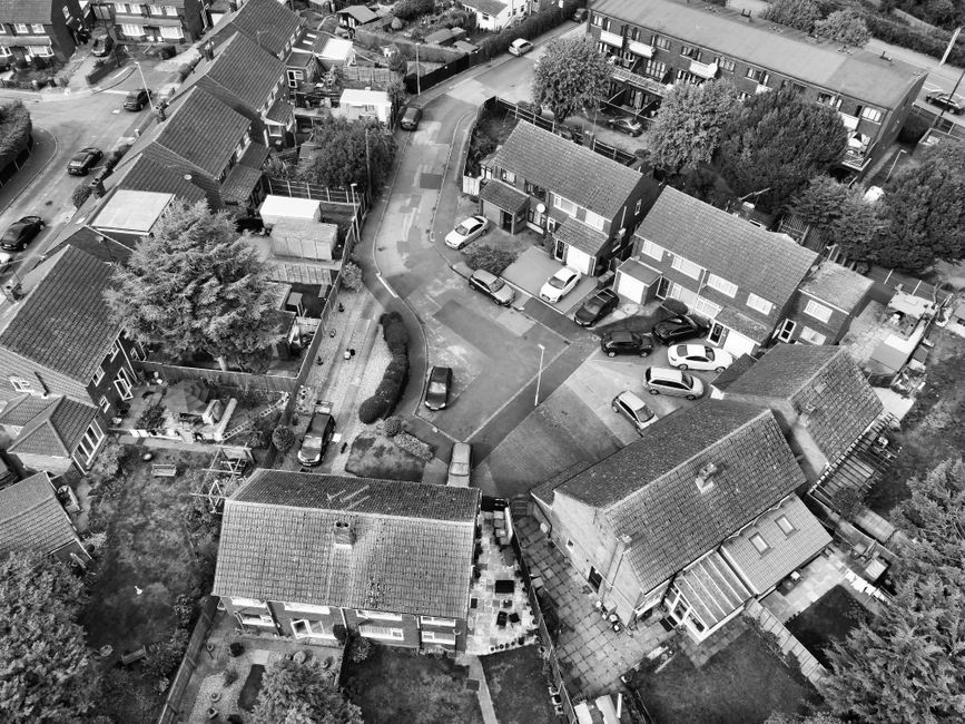 A black and white aerial photograph of a residential area in Upton Park, showing multiple terraced houses with pitched roofs and small front gardens. Several parked cars are visible along the narrow streets and driveways, with some vehicles positioned close to house entrances. In the central part of the image, a courtyard space is surrounded by houses, with a few cars parked and a curved driveway leading into the area. The surrounding environment includes trees, hedges, and fencing, indicating private gardens and outdoor spaces. The scene depicts a typical dense suburban neighbourhood suitable for home relocation or furniture transport, with visible brick pathways, roofing materials, and vehicle access points. This image captures the logistical context for house removals and packing and moving activities often handled by a company like Man with Van Upton Park, especially when addressing access problems on narrow streets during a move.