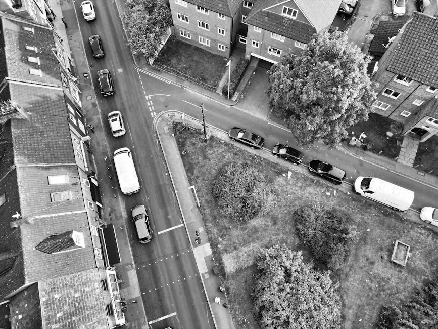 A black-and-white aerial photograph of a narrow residential street in Upton Park, showing multiple parked cars along the curb and a white van positioned on the paved roadway near a cluster of three trees and a small grassy area. The van is close to the entrance of a property, which has a short driveway and an intercom system visible on the wall. In the background, there are several multi-storey buildings with windows and pitched roofs. The scene captures the challenge of navigating tight street access for home relocation and furniture transport, with vehicles parked on both sides and the van being prepared for loading or unloading. Man with Van Upton Park offers house removals and packing services, with the image illustrating typical logistical considerations for moving in constrained urban environments.
