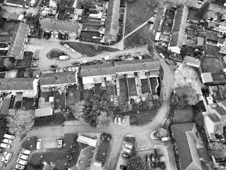 A black and white aerial photograph of a residential area in Upton Park, showing multiple terraced houses with pitched roofs and small front gardens. Several parked cars are visible along the narrow streets and driveways, with some vehicles positioned close to house entrances. In the central part of the image, a courtyard space is surrounded by houses, with a few cars parked and a curved driveway leading into the area. The surrounding environment includes trees, hedges, and fencing, indicating private gardens and outdoor spaces. The scene depicts a typical dense suburban neighbourhood suitable for home relocation or furniture transport, with visible brick pathways, roofing materials, and vehicle access points. This image captures the logistical context for house removals and packing and moving activities often handled by a company like Man with Van Upton Park, especially when addressing access problems on narrow streets during a move.