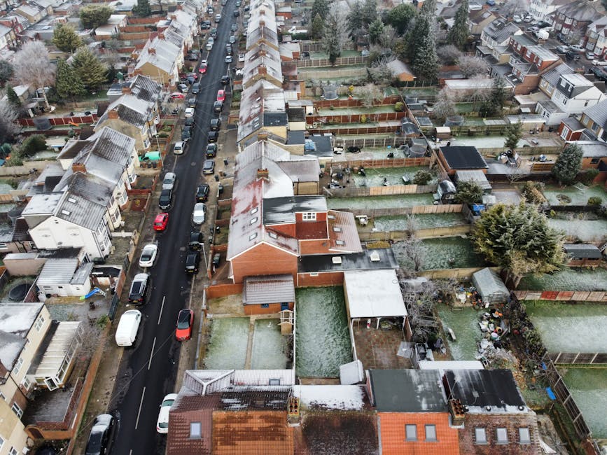 An aerial view of a residential street in Upton Park with houses and gardens, showing a line of parked cars along the road and a few driveways. Several backyards are visible, some enclosed with wooden fences, containing garden furniture, sheds, and trees. One garden has a small swimming pool covered with a green tarp, while another features outdoor seating and potted plants. The rooftops of the houses are partly covered with snow, and the scene is illuminated by natural daylight. The image illustrates a typical neighbourhood environment, relevant to house removals and moving services, as seen in the context of a home relocation or furniture transport process involving property access and outdoor space preparation, as managed by services like Man with Van Upton Park.