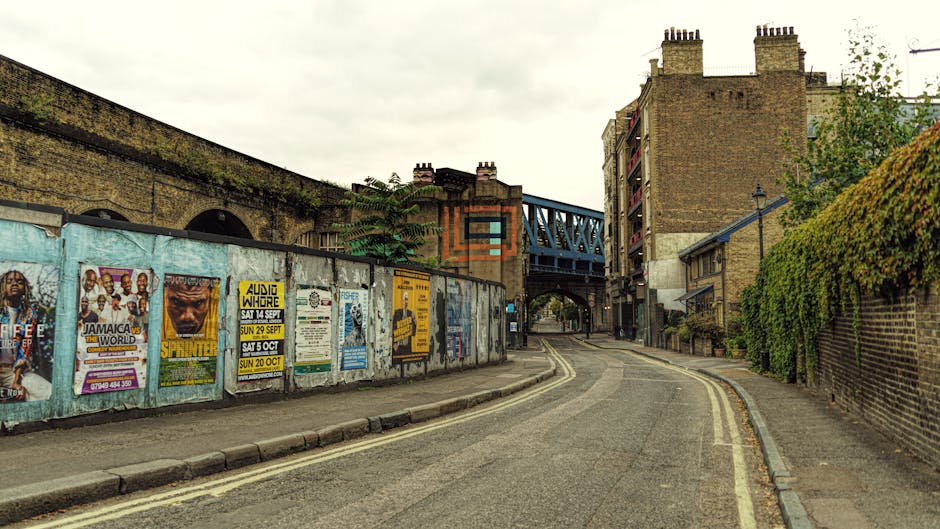 A long, straight urban street lined with mature trees showing green and yellow foliage on both sides, with a paved cycle lane or footpath on the left separated by a wooden post and grassy verge. On the right side, there are several parked cars facing away from the viewer, with some moving vehicles further down the road. The sky overhead is cloudy with dark, stormy clouds, suggesting a weather update. The scene is well-lit with natural light, and in the foreground, outside a property, there are numerous cardboard boxes, packing materials, and furniture blankets indicating a home relocation or furniture transport process. A person or team from Man with Van Upton Park may be involved in the loading or unloading activity across the curbside, with the focus on careful handling and safe transportation of household items during the moving process. This scene captures elements typical of house removals on Green Street and surrounding roads, illustrating the logistics and preparation involved in a professional moving service.