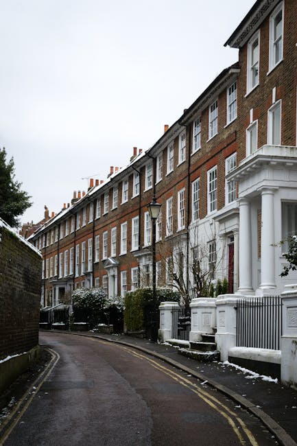 The image shows a narrow residential street in Upton Park during winter, with an overcast sky and a light dusting of snow on the pavement and low garden walls. Traditional terraced houses made of red brick with white architectural details line the street, featuring large sash windows and decorative columns at the entrances. In the foreground, a black van is parked on the pavement, adjacent to the base of a lamppost. To the right, a white-painted gated porch with steps leads up to a house entrance. The street is quiet with no visible pedestrians or moving vehicles, emphasizing a calm, urban environment suitable for house removals. Man with Van Upton Park offers professional local removal services, including furniture transport and packing, suitable for tight-access flats like those depicted in this typical London residential setting.