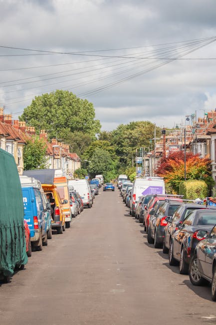 A residential street scene during daylight hours showing parked cars lining both sides of the narrow tarmac road, with a mix of small hatchbacks, sedans, and vans. Some of the vans display company branding related to removals and home relocation services, indicating presence of professional moving activities. The pavement borders the houses, which are two-storey terraced properties with brick facades, pitched roofs, and small front gardens or steps leading to front doors. The street is flanked by green trees and well-maintained shrubs, with overhead power lines visible against a partly cloudy sky, suggesting typical urban residential surroundings. This setting illustrates an environment where house removals and furniture transport might be taking place, supported by the presence of specialised moving vehicles and equipment visible in the scene, such as blankets or straps used during the loading and unloading process, aligned with the context of packing and home relocation in the Upton Park area, as outlined in the Green Street Upton Park moving guide for tight-access flats. Man with Van Upton Park's services are indirectly referenced through the setting's typical professional moving environment.