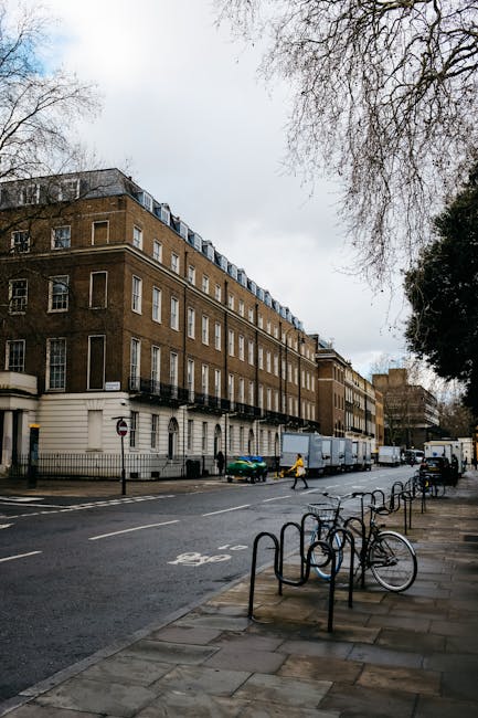 The image shows a narrow residential street in Upton Park during winter, with an overcast sky and a light dusting of snow on the pavement and low garden walls. Traditional terraced houses made of red brick with white architectural details line the street, featuring large sash windows and decorative columns at the entrances. In the foreground, a black van is parked on the pavement, adjacent to the base of a lamppost. To the right, a white-painted gated porch with steps leads up to a house entrance. The street is quiet with no visible pedestrians or moving vehicles, emphasizing a calm, urban environment suitable for house removals. Man with Van Upton Park offers professional local removal services, including furniture transport and packing, suitable for tight-access flats like those depicted in this typical London residential setting.