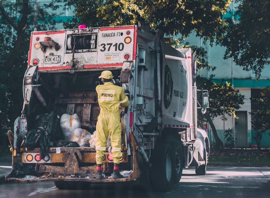 A waste collection truck with a white cab and red rear section is parked on a street, with the rear loading compartment open. Inside, there are multiple black garbage bags filled with waste, some tied with ribbons or string, and a few clear plastic bags containing recyclables or packaging materials. A worker in a yellow high-visibility uniform, including a helmet and reflective strips, is standing at the back of the truck, using a tool or controlling the loading process. The truck is positioned near a sidewalk, with some greenery and trees visible in the background, and sunlight filtering through the leaves. The surroundings include a modern building with blue and white tones, indicating an urban environment. This scene reflects proper waste management procedures, which are an important aspect of house removals and property clearing, handled by companies like Man with Van Upton Park, ensuring compliance with legal obligations for waste disposal during home relocations.