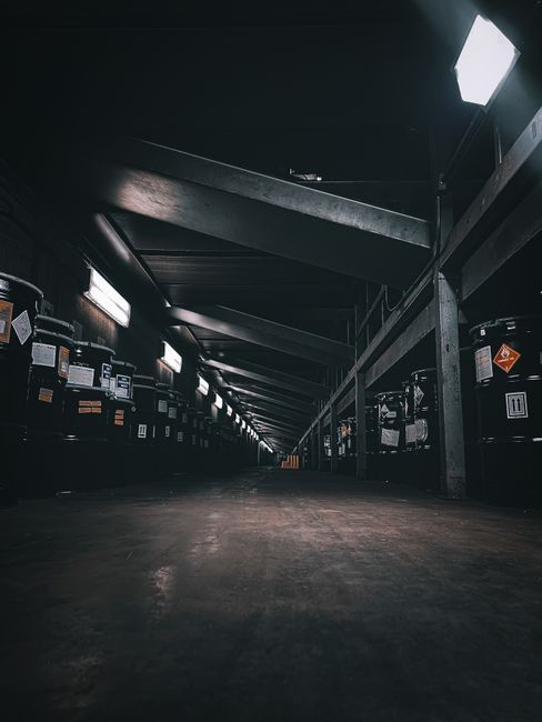 A low-angle view inside a dimly lit storage or warehouse area with dark metal beams and shelving units lining both sides. Several large black plastic containers with labels and hazard symbols are stacked along the shelves, with some boxes visible among them. The floor appears to be wooden or concrete, showing signs of wear. The space is illuminated by small overhead lights, creating a stark contrast between the dark environment and the illuminated areas. This setting reflects the storage aspect of household items before or during a home relocation, highlighting the packing and safety measures involved. Occasionally, [COMPANY_NAME] provides professional removals and furniture transport services, ensuring safe handling of household belongings such as furniture, boxes, and appliances during the loading process. The image captures the organized storage environment where packed items are likely prepared for transportation, supporting the overall theme of packing, moving, and logistics associated with house removals in the context of licensed waste carriers in Upton Park.