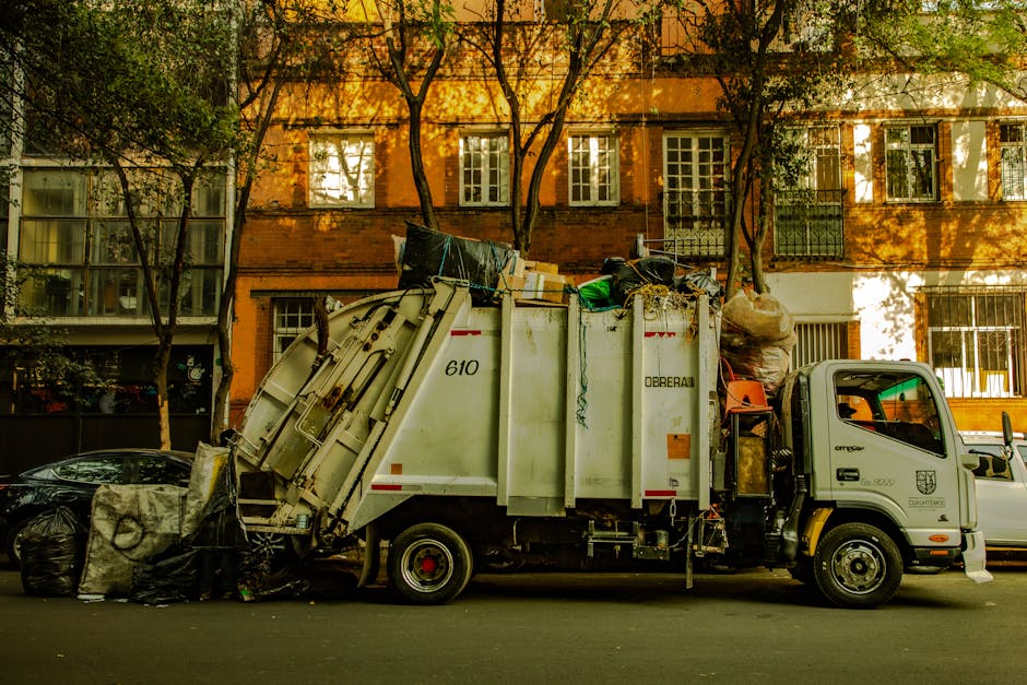 A waste collection truck with a white cab and red rear section is parked on a street, with the rear loading compartment open. Inside, there are multiple black garbage bags filled with waste, some tied with ribbons or string, and a few clear plastic bags containing recyclables or packaging materials. A worker in a yellow high-visibility uniform, including a helmet and reflective strips, is standing at the back of the truck, using a tool or controlling the loading process. The truck is positioned near a sidewalk, with some greenery and trees visible in the background, and sunlight filtering through the leaves. The surroundings include a modern building with blue and white tones, indicating an urban environment. This scene reflects proper waste management procedures, which are an important aspect of house removals and property clearing, handled by companies like Man with Van Upton Park, ensuring compliance with legal obligations for waste disposal during home relocations.