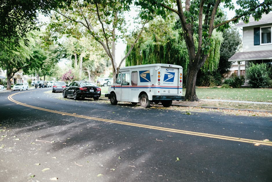 A United States Postal Service delivery truck, painted white with blue and red accents, parked partially on the sidewalk and street near a residential area during daytime. The truck is positioned beside a large tree with green foliage, and behind it, several cars are parked along the curved, asphalt road with yellow double lines. The sidewalk is adjacent to a lawn with bushes and a house with a white façade, dark roof, and visible windows. The scene is well-lit with natural daylight, and the area appears calm and quiet. This image may relate to house removals or moving services by showing parking and loading considerations for a delivery vehicle in a suburban environment, relevant to furniture transport and home relocation logistics involving [COMPANY_NAME] at [PAGE_URL].