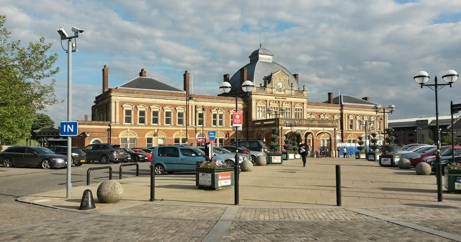 A view of Upton Park railway station with a large, historic brick building featuring decorative stonework, arched windows, and a clock on the central facade. The foreground includes a paved area with black metal barriers, lamp posts with spherical lamps, and large spherical stone bollards. Several parked cars occupy the parking area adjacent to the station, with some moving vehicles visible. A sign indicating 'IN' is mounted on a pole near the pavement. The scene is well-lit under a partly cloudy sky, providing natural daytime illumination. This setting is suitable for house removals or furniture transport services, such as those offered by Man with Van Upton Park, which may involve loading furniture, boxes, and appliances for home relocation within or around the area. The environment shows activity related to moving logistics with a focus on parking and access points for moving vans in the vicinity of the station.
