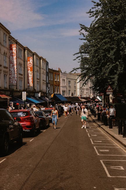 The image depicts a busy street scene outside Queen's Market in Upton Park on a clear day with blue skies and some scattered clouds. On the left side, there are several multi-story buildings with retail storefronts at ground level, some with large vertical banners displaying promotional graphics. The street is lined with parked cars, including a red vehicle in the foreground, and several pedestrians are walking along the pavement, some carrying shopping bags. To the right, there is a row of black bollards separating the sidewalk from the parking area. A large tree with leafy branches extends over part of the scene, providing some shade. The overall setting suggests a lively market environment, with the context aligning with house removals and packing activities, as individuals and vehicles are preparing for or conducting a home relocation process. Man with Van Upton Park may utilize such streets for loading or unloading furniture during a furniture transport or moving service, evidenced by the organized arrangement of parked vehicles and pedestrian activity.