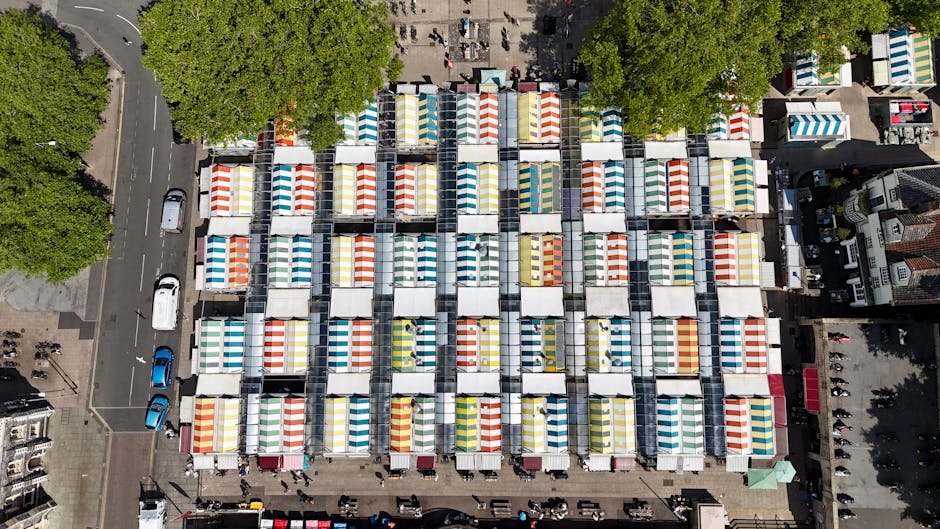 An aerial view of a large outdoor market with multiple rows of colorful striped canopies arranged on rectangular stalls, situated on a paved area surrounded by trees and adjacent buildings. The market is densely populated with pedestrians walking between the stalls. A few parked cars are visible along the street borders, and the setup appears organized for a community event or local fair. This scene illustrates the typical environment for home relocation or packing and moving services provided by Man with Van Upton Park, capturing the logistical aspects of moving day preparations and the importance of careful planning during furniture transport and equipment loading processes.