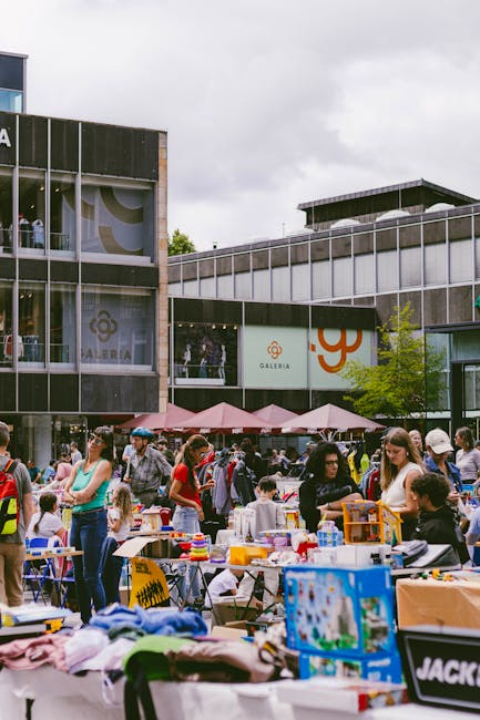 The image depicts a busy street scene outside Queen's Market in Upton Park on a clear day with blue skies and some scattered clouds. On the left side, there are several multi-story buildings with retail storefronts at ground level, some with large vertical banners displaying promotional graphics. The street is lined with parked cars, including a red vehicle in the foreground, and several pedestrians are walking along the pavement, some carrying shopping bags. To the right, there is a row of black bollards separating the sidewalk from the parking area. A large tree with leafy branches extends over part of the scene, providing some shade. The overall setting suggests a lively market environment, with the context aligning with house removals and packing activities, as individuals and vehicles are preparing for or conducting a home relocation process. Man with Van Upton Park may utilize such streets for loading or unloading furniture during a furniture transport or moving service, evidenced by the organized arrangement of parked vehicles and pedestrian activity.