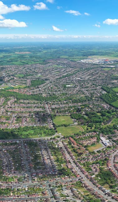 An aerial view of a suburban area showing residential streets lined with houses, surrounded by green spaces and parks, with a large commercial or industrial area in the distance beneath a bright blue sky with scattered clouds. The image captures the urban layout during daylight hours, highlighting the organization of housing and open areas, which is relevant to house removals and moving services offered by Man with Van Upton Park. This perspective emphasizes the logistics involved in home relocation projects, including the planning of transport routes and loading processes for furniture and belongings, set against the backdrop of a typical UK neighborhood.