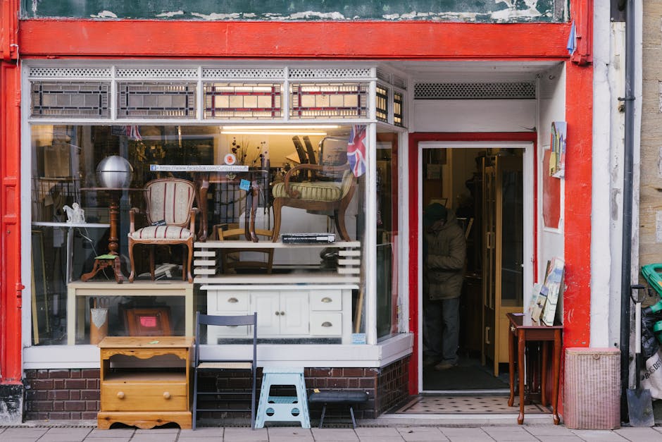 The storefront of a second-hand furniture shop, with large glass windows displaying various antique and vintage furniture pieces, including armchairs, a wooden side table, and a white cabinet, some de