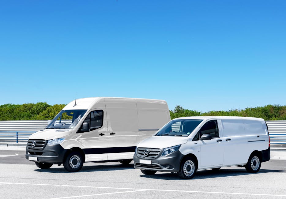 Two white cargo vans parked side by side on a paved surface next to a metal guardrail, with a clear blue sky above and green trees in the background. The larger van on the left features a high roof an