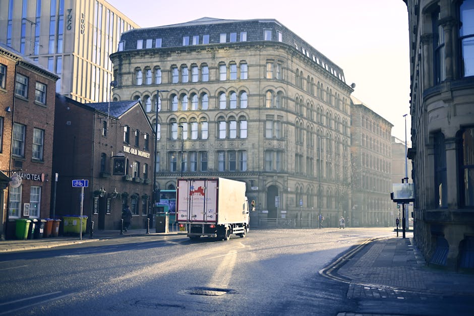 A white moving truck with red branding is positioned on a city street, partially visible in the center of the image, loaded with cardboard boxes sealed with packing tape and wrapped with plastic stretch film, which are stacked on wooden dollies for transport. The truck is parked next to a sidewalk, with several large, multi-storey historic buildings featuring arched windows and stone facades on the opposite side of the street, typical of an urban environment suitable for house removals and furniture transport. Nearby, on the left, there are smaller brick buildings, and leafless trees and bins are visible along the pavement. The scene is illuminated by natural daylight, creating long shadows on the asphalt, indicating a clear day. The overall setting suggests an ongoing or planned home relocation, with the loading process of furniture and household items into the vehicle, supported indirectly by the logistics services offered by Man with Van Upton Park for prompt, door-to-door removals in Upton Park.